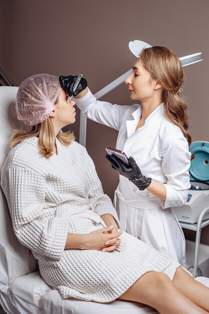 Il cliente in berretto e vestaglia riceve segni sul viso con una penna nelle mani di un cosmetologo esperto per fornire servizi di cosmetologia