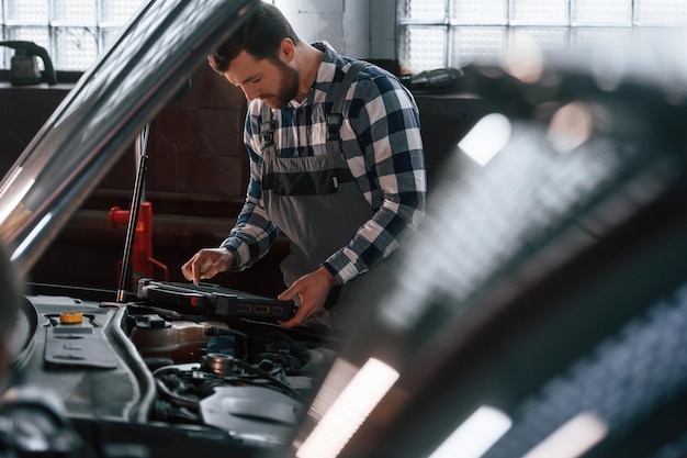 Il cofano dell'auto è aperto Utilizzando il tablet L'uomo in uniforme sta lavorando nel salone dell'auto