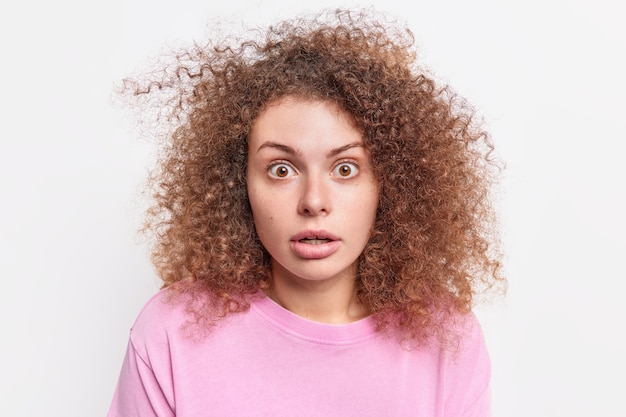 Il colpo alla testa della donna stordita con i capelli folti ricci fissa con incredulità reagisce a notizie incredibili indossa abiti casual isolati sul muro bianco. Concetto di reazione ed emozioni umane