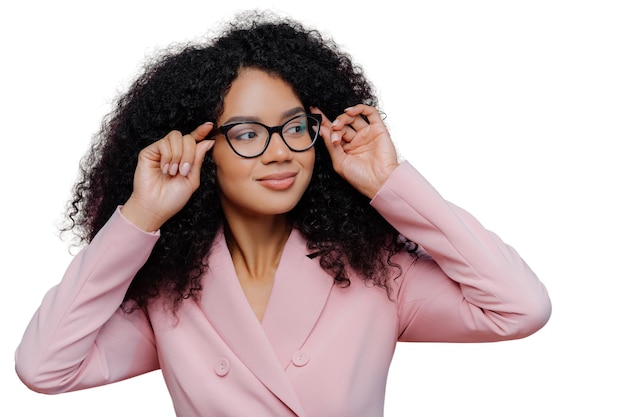 Il colpo alla testa di un capo femminile attento tiene le mani sulla montatura degli occhiali guarda pensieroso lontano indossa un abito formale roseo ha un'acconciatura afro riccia posa su sfondo viola persone etnia aziendale