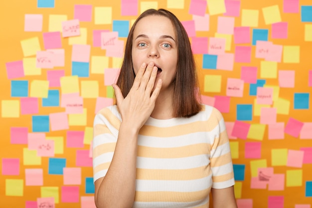 Il colpo orizzontale di una donna caucasica sorpresa che indossa una maglietta a righe vede qualcosa di sorprendente che dice wow che copre la bocca con la mano in piedi contro il muro giallo coperto di adesivi multicolori