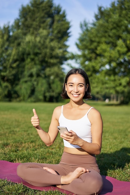 Il colpo verticale della donna asiatica mostra i pollici in su raccomandando l'app di meditazione online per l'allenamento yoga