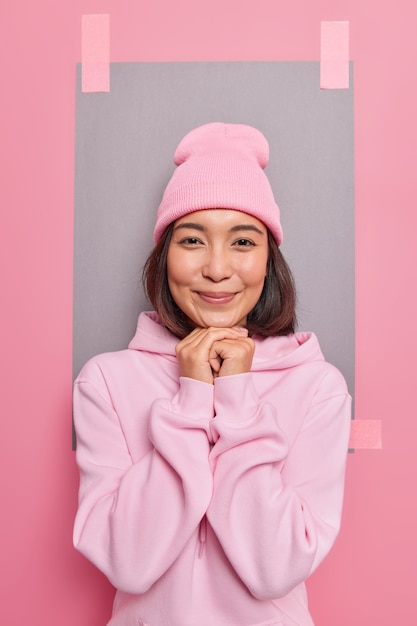 Il colpo verticale di bella donna asiatica con i capelli scuri tiene le mani sotto il mento sorride indossa delicatamente il cappello e la felpa con cappuccio guarda direttamente le pose della fotocamera contro il foglio bianco intonacato al muro