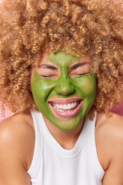 Il colpo verticale di una donna dai capelli ricci felice chiude gli occhi sorride ampiamente mostra i denti bianchi applica una maschera di bellezza verde per il trattamento della pelle indossa una maglietta bianca casual Cura del viso e concetto di spa benessere