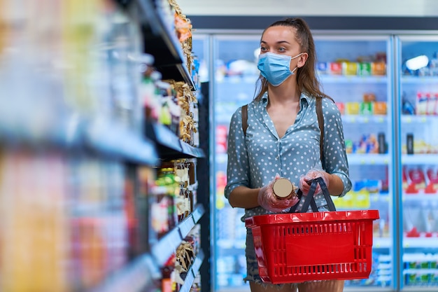 Il compratore della donna in una maschera protettiva con il cestino della spesa sceglie l'alimento dei prodotti sugli scaffali in un negozio di alimentari