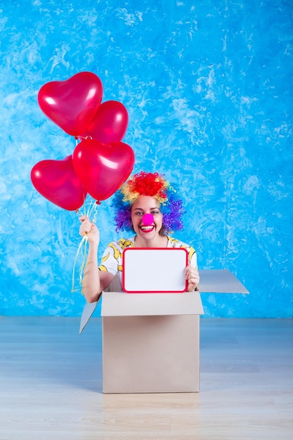 Il concetto del giorno dei pazzi del 1° aprile La ragazza clown divertente che ride si siede in una scatola di cartone con palloncini di elio e tavola bianca in mano Regalo di compleanno