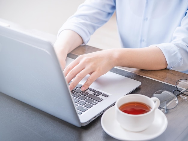 Il concetto di business donna in camicia blu digitando sul computer con caffè sul tavolo dell'ufficio retroilluminazione effetto abbagliamento del sole primo piano vista laterale spazio di copia