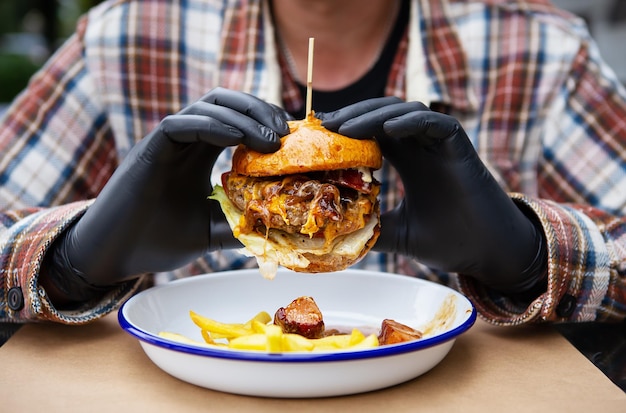 Il concetto di fast food e cibo da asporto Un uomo in guanti di lattice neri tiene in mano un hamburger succoso si trova vicino a patatine fritte su una piastra di metallo insieme a salsa di formaggio