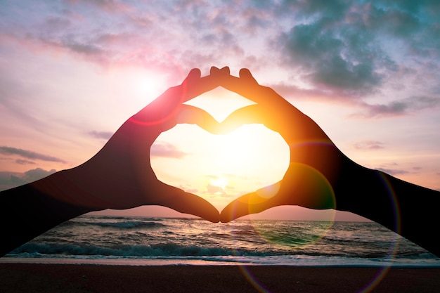Il concetto di giorno di San Valentino, la mano della siluetta fa la forma del cuore con il cielo e la luce del sole di vista sul mare.