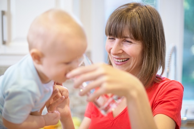 Il concetto di maternità, tata, infanzia e infanzia. Tiro al coperto in cucina. Due donne e un bambino tra le braccia, al bambino viene data acqua, una famiglia felice