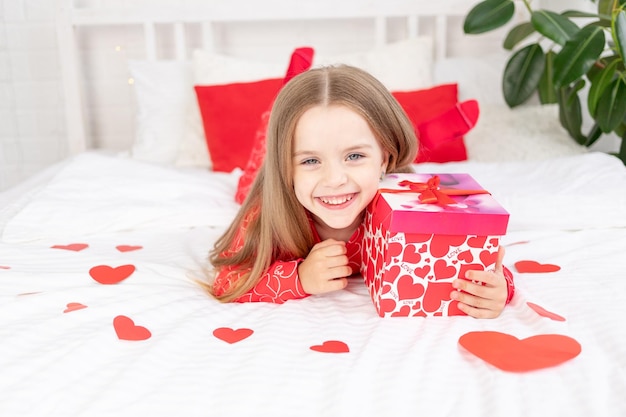 Il concetto di San Valentino una bambina carina è seduta sul letto a casa in pigiama rosso e tiene un regalo tra le mani e sorride o ride di felicità congratulandosi per la vacanza