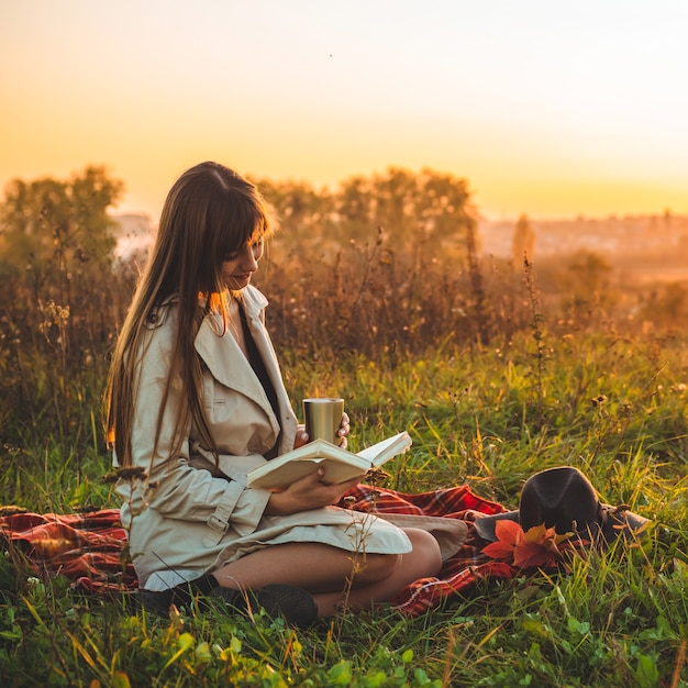 Il concetto di stile di vita e attività ricreative all'aperto in famiglia in autunno. donna con cappello legge libri su plaid con una tazza termica. Autunno. Tramonto. Accogliente