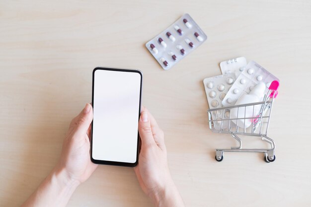 Il concetto di un'applicazione mobile per l'acquisto a distanza di medicinali Farmacia online Uno smartphone con uno schermo bianco in mani femminili e un mini carrello con blister per tablet