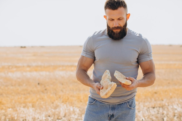 Il contadino felice dell'uomo barbuto in jeans tiene il pane fresco e profumato strappandolo a metà e in piedi in un campo soleggiato intorno alle balle