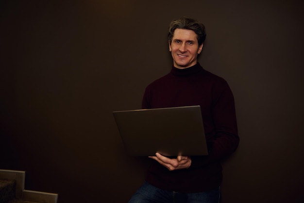 Il copywriter imprenditore europeo maturo bello lavora online su laptop in piedi contro una parete scura illuminata con luce fioca in ufficio