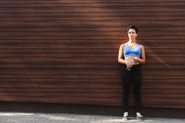 Il corridore della giovane donna sta facendo una pausa, bevendo acqua mentre fa jogging in città, appoggiandosi al muro di legno scuro, copia spazio