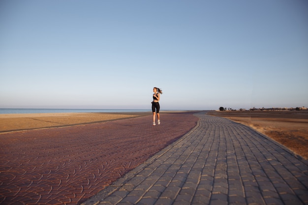 Il corridore della ragazza in una tuta nera e scarpe da ginnastica corre in riva al mare durante il tramonto