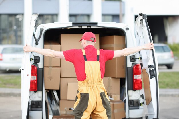 Il corriere maschio in uniforme apre il bagagliaio di un minivan pieno di scatole di cartone