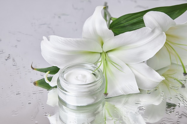 Il cosmetico crema barattolo di bellezza con fiori di giglio si trova sul tavolo, prodotto crema barattolo su specchio bagnato inumidito. Cosmetici in crema per vasetti di fiori naturali per la cura della pelle delle mani. Pelle liscia e di bellezza