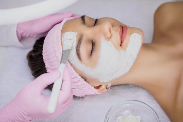 Il cosmetologo applica una maschera all'argilla bianca sul viso della ragazza con un pennello bianco