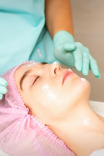 Il cosmetologo con le mani guantate applica una maschera idratante con crema peeling sul viso femminile Trattamento di cosmetologia facciale Procedure per la cura del viso