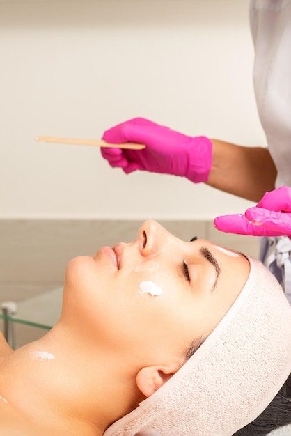 Il cosmetologo con le mani guantate applica una maschera idratante con crema peeling sul viso femminile Trattamento di cosmetologia facciale Procedure per la cura del viso