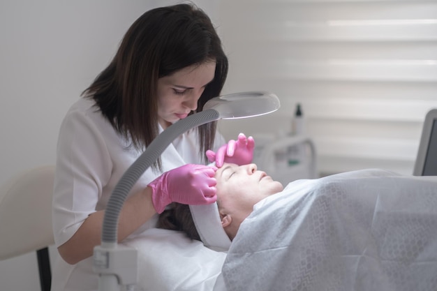 Il cosmetologo esamina la pelle del cliente attraverso la lampada d'ingrandimento durante un trattamento viso professionale