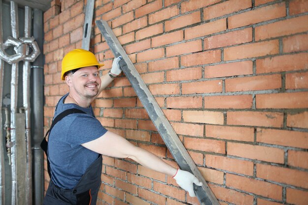 Il costruttore felice tiene i dettagli in metallo vicino al muro di mattoni in cemento rosso nella stanza vuota