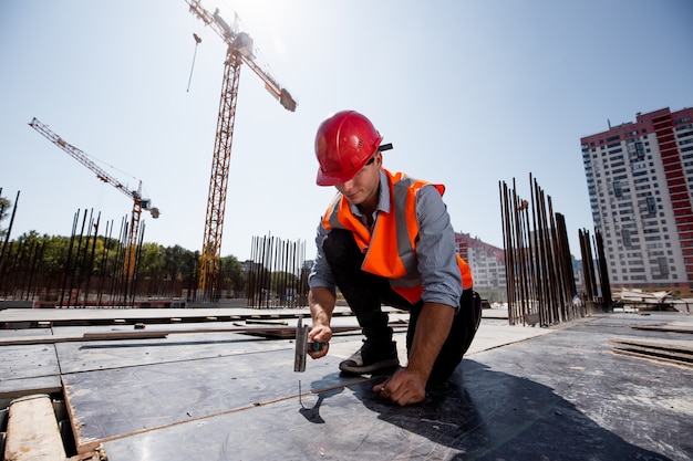 Il costruttore vestito con giubbotto da lavoro arancione e casco martella un chiodo sul cantiere aperto.