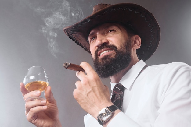 Il cowboy barbuto fiducioso sta fumando sigari e bevendo alcolici su sfondo grigio.