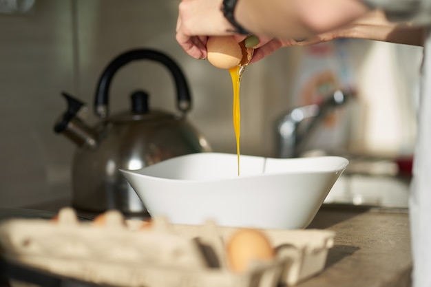 Il cracking caucasico della donna apre un uovo durante la cottura della prima colazione. Chiudi le mani