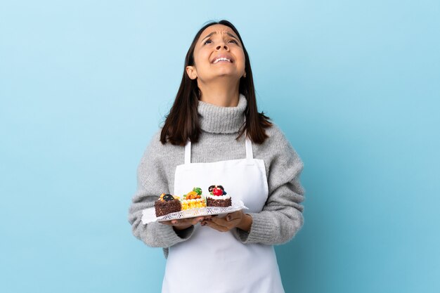 Il cuoco unico di pasticceria che tiene una grande torta sopra la parete blu isolata tiene insieme la palma. La persona chiede qualcosa