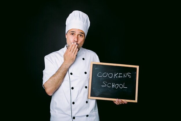 Il cuoco vestito con una giacca e un cappello bianco si porta la mano alla bocca e tiene una lavagna che recita: Scuola di cucina su sfondo nero. Ristorante, cibo e concetto di scuola di cucina.