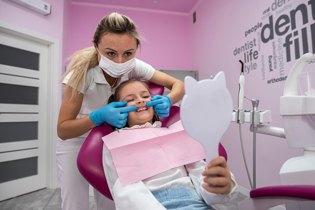 Il dentista femminile mostra i denti trattati alla bambina paziente davanti allo specchio odontoiatria per bambini bellissimo piccolo sorriso specchio nelle mani