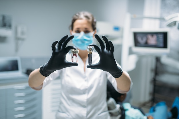 Il dentista femminile mostra il perno del dente e il modello, la clinica odontoiatrica, l'odontoiatria protesica professionale, l'armadietto per stomatologia, l'odontoiatria