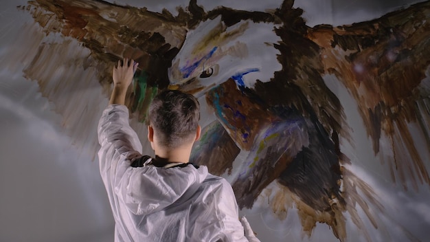 Il designer artista disegna un'aquila sul muro Il decoratore artigiano dipinge l'immagine con colore ad olio acrilico Pittore pittore vestito con vernice per interni