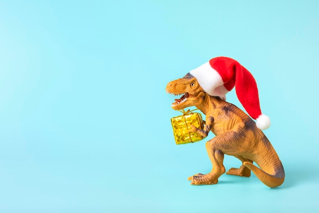 Il dinosauro Rex con il cappello rosso di Babbo Natale tiene una scatola regalo dorata tra le zampe su sfondo blu