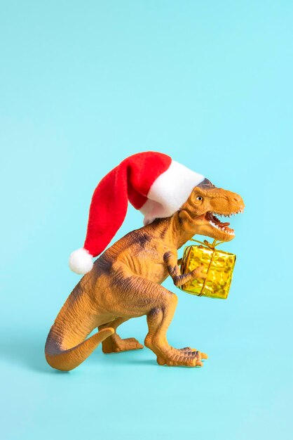 Il dinosauro Rex con il cappello rosso di Babbo Natale tiene una scatola regalo dorata tra le zampe su sfondo blu