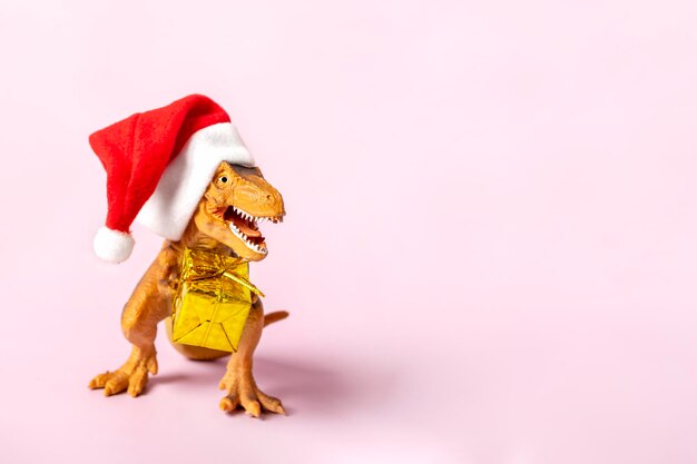 Il dinosauro Rex con il cappello rosso di Babbo Natale tiene una scatola regalo dorata tra le zampe su sfondo rosa Biglietto di auguri di Capodanno o vigilia di Natale Art Idea creativa per il concetto di Buon Natale