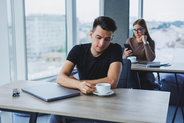 Il dipendente del manager si siede a un tavolo di legno e lavora su un laptop C'è un caffè e un laptop sul tavolo Ufficio con grandi finestre