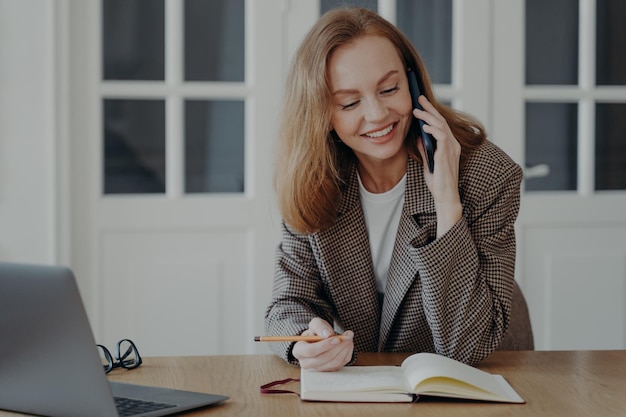 Il dipendente femminile risponde alla chiamata alla scrivania con il computer portatile che parla al telefono che consulta il cliente che prende appunti