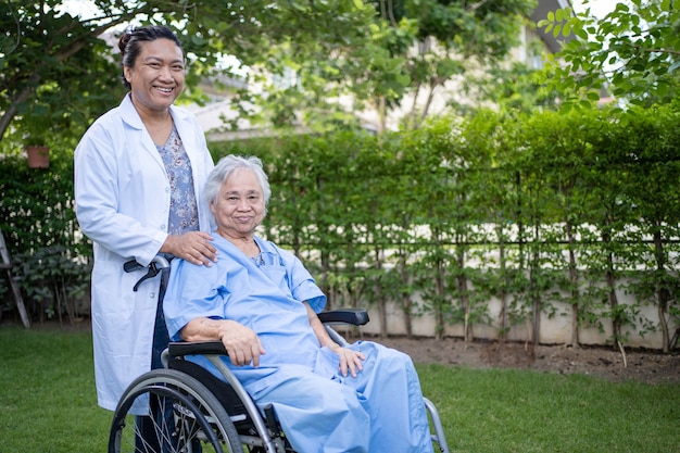 Il dottore aiuta e cura una paziente asiatica anziana seduta su una sedia a rotelle al parco