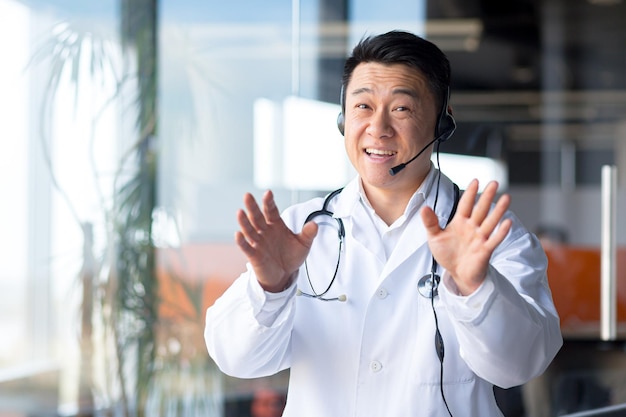 Il dottore felice sorride e guarda la telecamera, l'asiatico annuncia buone notizie al paziente, usa una videochiamata e un incontro online con le cuffie con i colleghi, gesticola divertente con le mani, vista dalla webcam