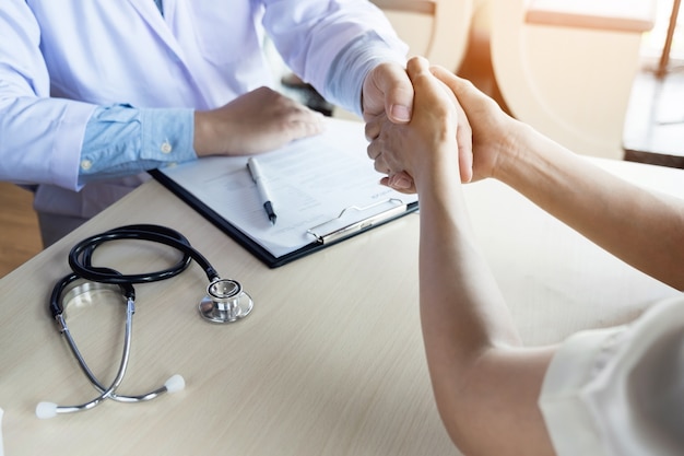 Il dottore stringe la mano all&#39;ufficio medico con paziente, indossa occhiali, stetoscopio e cappotto di laboratorio.