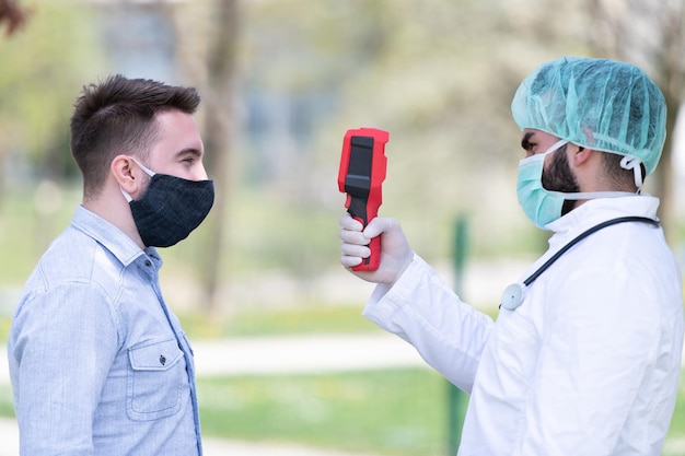 Il dottore usa la pistola del termometro a infrarossi sulla fronte per controllare la temperatura corporea per i sintomi del virus Covid-19 con l'abito isolante o tute protettive e maschere chirurgiche fuori nel parco
