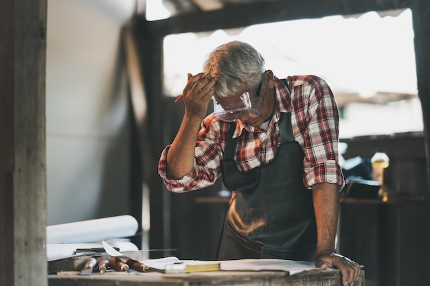 Il falegname senior usa la mano sopra la testa pensando o serio o triste o depressione per progetti di lavoro su macchine per la lavorazione del legno in falegnameria l'uomo lavora in una falegnameria