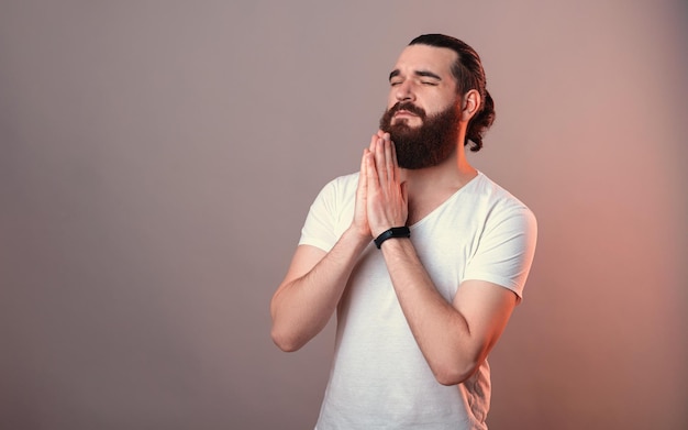 Il fedele uomo barbuto con gli occhi chiusi tiene le mani per una preghiera Studio girato con luce rossa su sfondo grigio