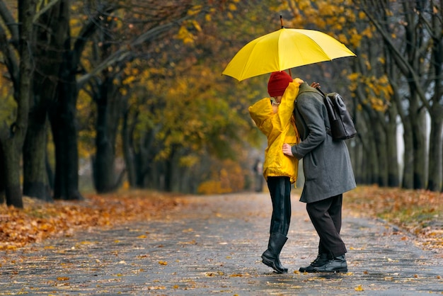 Il figlio sta abbracciando sua madre che cammina nel parco autunnale sotto la pioggia con un grande ombrello giallo Giornata piovosa