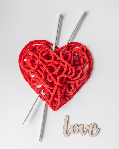 Il filato per maglieria rosso con gli aghi e il segno di legno AMANO sulla tavola bianca. A forma di cuore. Concetto minimo di San Valentino.