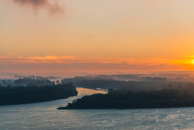 Il fiume scorre lungo la riva con la foresta sotto la nebbia. Il bagliore arancione nel cielo dell'alba si riflette sull'acqua.